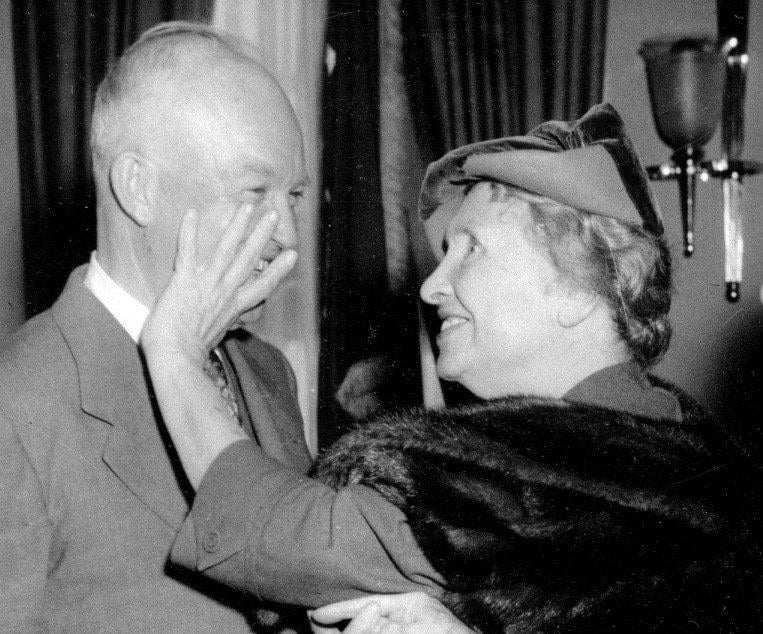 A woman wearing a fur stole and hat gently touches the face of an older man in a suit; both are smiling warmly at each other in an indoor setting with curtains and a wall lamp.
