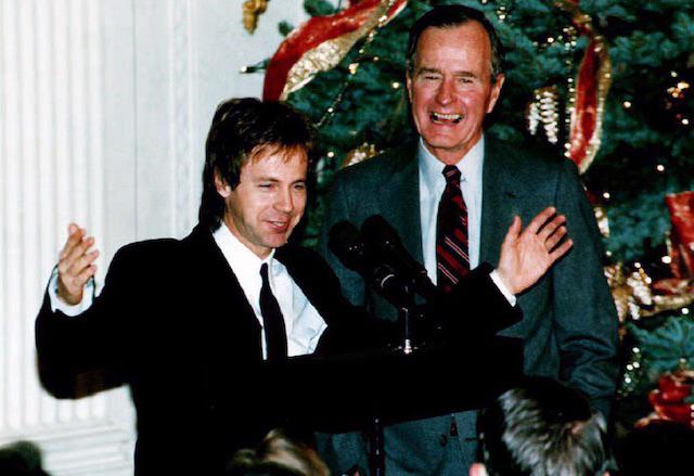 Two men in suits stand at a podium, smiling and speaking. There is a decorated Christmas tree in the background, suggesting a holiday event. One man gestures with open arms while the other stands behind him, laughing.