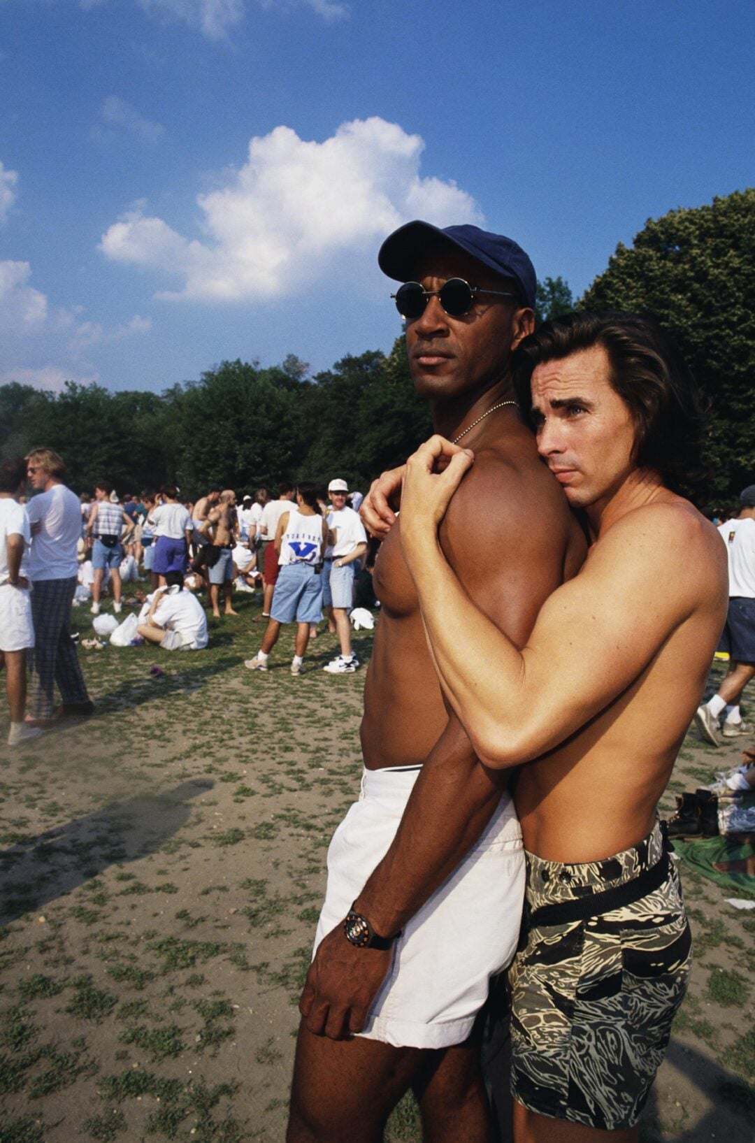 Two shirtless men stand close together outdoors; one hugs the other from behind. They are surrounded by a crowd of casually dressed people in a sunny park with trees in the background.