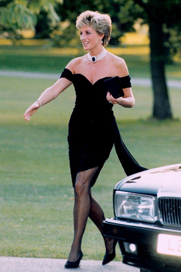 A woman in a black off-the-shoulder dress, black heels, and a white choker necklace walks on grass near a parked car, holding a black clutch. Trees and greenery are visible in the background.