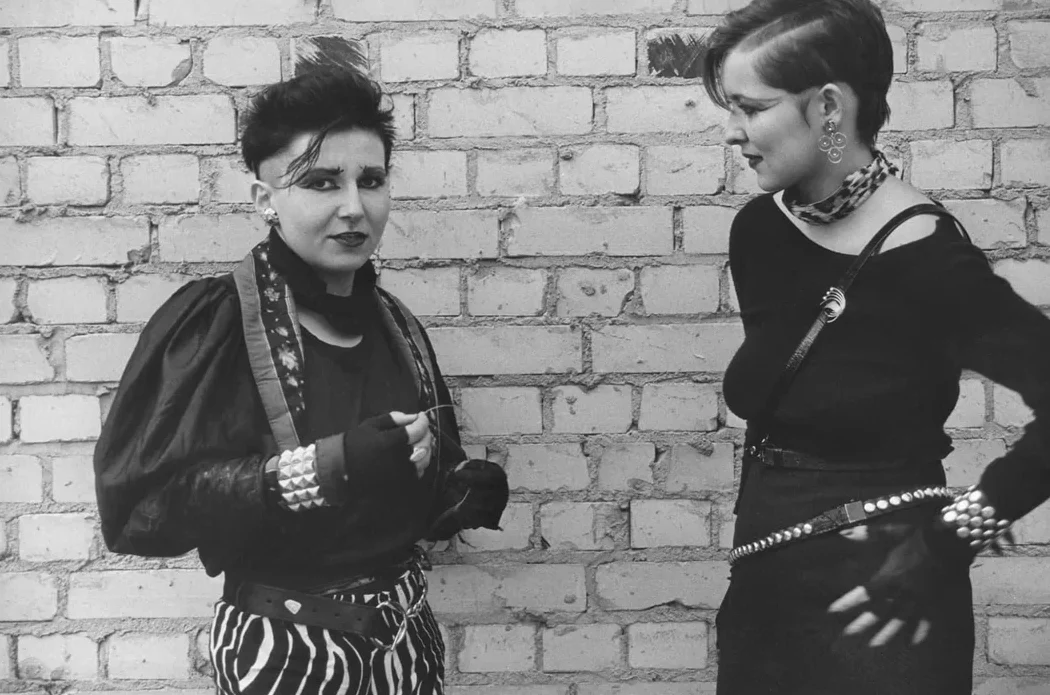 Two people with punk-style clothing and short, edgy haircuts stand in front of a brick wall. One wears striped pants and studded bracelets; the other has a choker, earrings, and a studded belt. Both have expressive, confident poses.