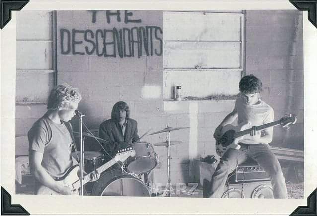 A black-and-white photo shows three young musicians performing in a garage. The wall behind them has "THE DESCENDANTS" painted on it. One plays guitar, one is on drums, and one is playing bass while sitting on an amp.