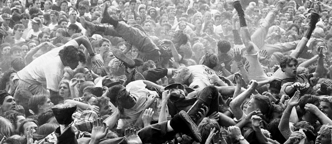 A dense crowd at a concert with several people crowd surfing above raised hands, creating a chaotic and energetic atmosphere. The image is black and white.