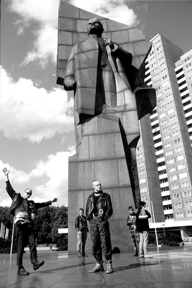 A group of young people in punk clothing stand near a large statue of Lenin, with modern high-rise buildings and a partly cloudy sky in the background. One person raises an arm energetically.