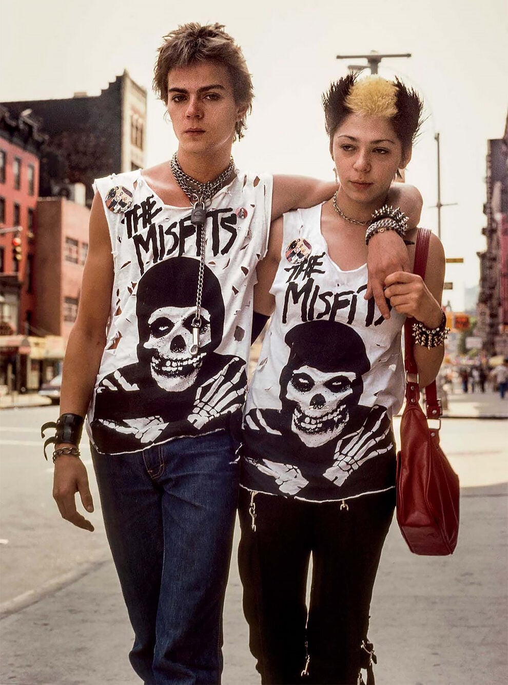 Two young people with punk hairstyles and accessories walk arm in arm down a city street, both wearing ripped "The Misfits" T-shirts, black pants, and bold jewelry. Urban buildings are visible in the background.