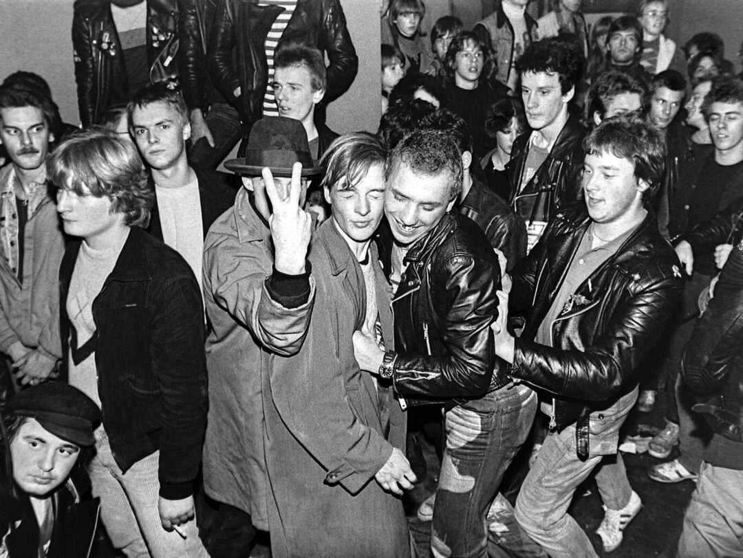 A black-and-white photo of a crowded punk concert, showing people in leather jackets. One person in the center holds up two fingers in a V sign, while others smile, laugh, or look toward the camera. The scene is lively and energetic.
