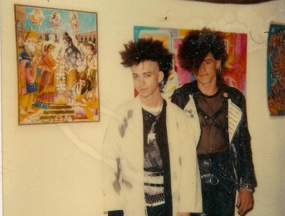 Two people with spiked, voluminous hair and punk-style outfits stand indoors near colorful, framed artworks on the wall, including one with Hindu deities. The photo has a vintage, slightly faded look.
