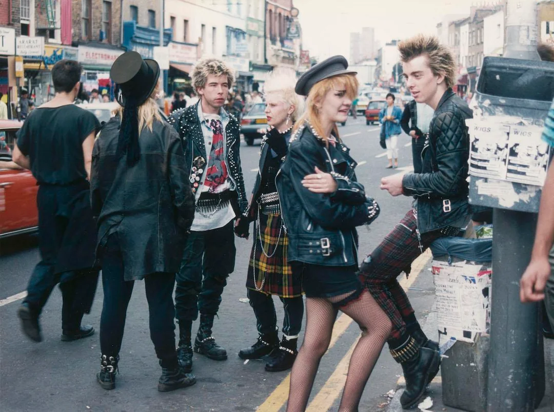 A group of punk youth in leather jackets and plaid clothing stand and chat on a busy city street lined with shops and posters, embodying 1980s punk fashion and attitude.