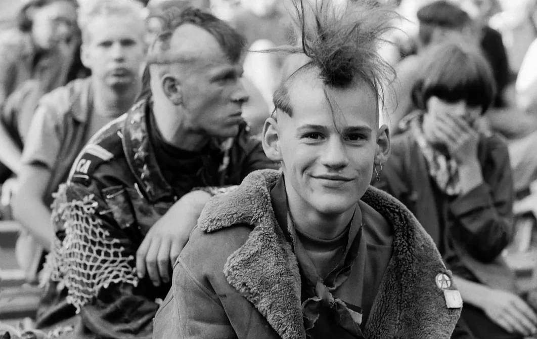 A young man with a spiked mohawk and a fur-collared jacket smiles at the camera. Behind him, others with punk hairstyles and eclectic clothing sit together in an outdoor setting.