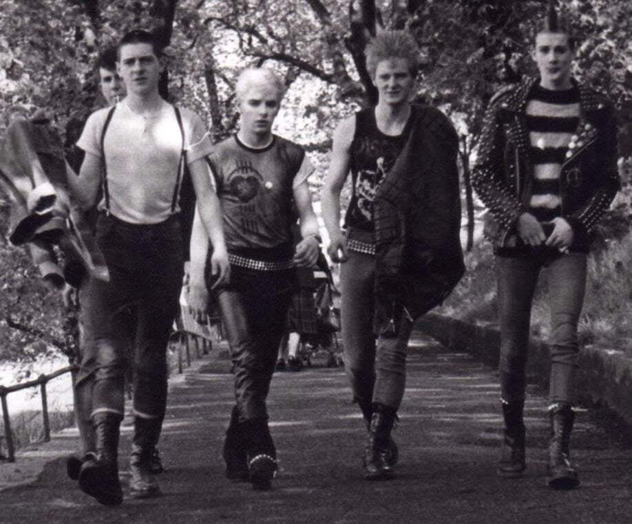 Four young men dressed in punk attire, including boots, suspenders, studded belts, and leather jackets, walk confidently down a tree-lined path. The image is black and white, giving it a vintage feel.