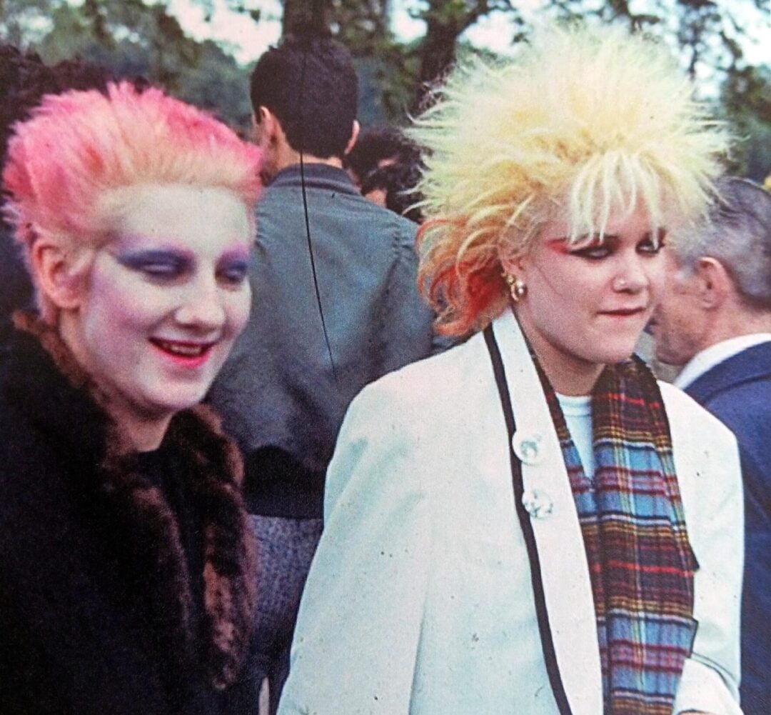 Two women with brightly colored spiked hair and bold makeup are outdoors. One wears a fur-collared coat, the other a white jacket and plaid scarf. Both have a punk style, with people and trees visible in the background.