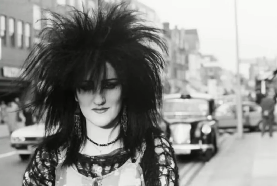 A woman with voluminous, teased black hair and dark makeup stands on a city street. Vintage cars and buildings are visible in the blurred background, giving the scene a retro atmosphere.