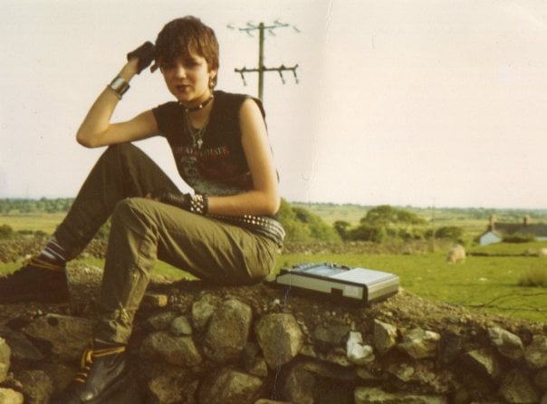 A person with short hair and punk-style clothing sits on a stone wall in the countryside, holding their hand to their forehead. A portable cassette player sits next to them. Fields and power lines are visible in the background.