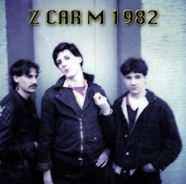 Three young men stand closely together in front of a wall, dressed in casual jackets. Bold yellow text above them reads "Z CAR M 1982." The image has a vintage, slightly blurred look.