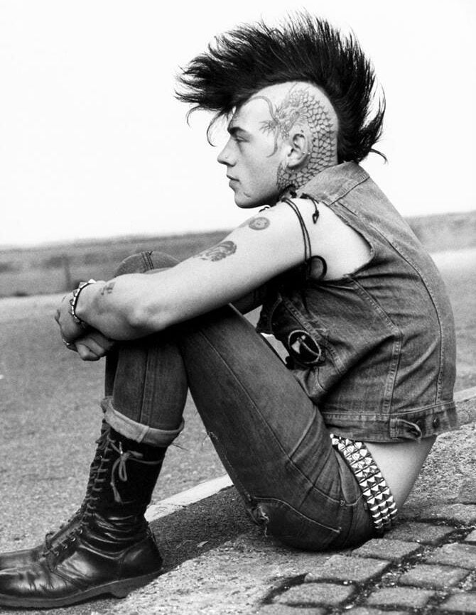 A person with a tall mohawk, head tattoos, and piercings sits on a curb wearing a sleeveless denim vest, tight jeans, studded belt, and lace-up boots, looking pensively to the side.