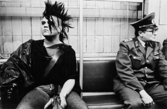 A person with a spiked mohawk and punk attire sits on a subway bench beside a uniformed officer; both look in opposite directions, creating a striking contrast in appearance and attitude.