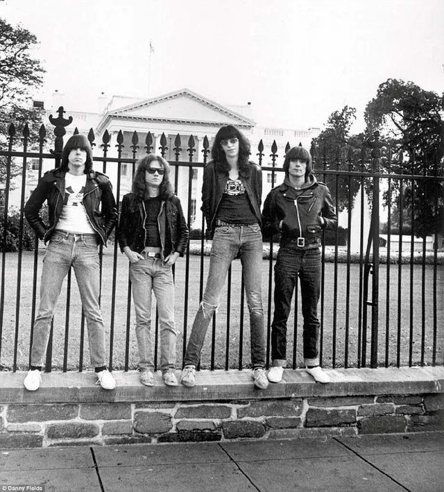 Four men in leather jackets, jeans, and sunglasses stand in front of a tall iron fence with the White House visible in the background. The photo is black and white and appears to be from the 1970s or 1980s.