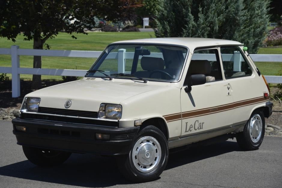 A vintage cream-colored hatchback car with "Le Car" written on the side is parked on a street near a white fence and greenery. The car has two doors and classic styling from the late 1970s or early 1980s.