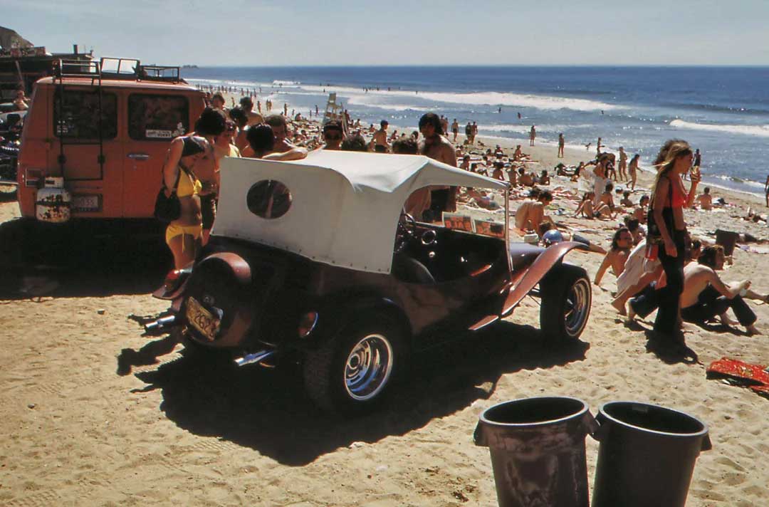 A busy beach scene with people sunbathing and playing near the ocean. In the foreground, an orange van and a red dune buggy are parked on the sand beside two trash cans. The weather is sunny and clear.