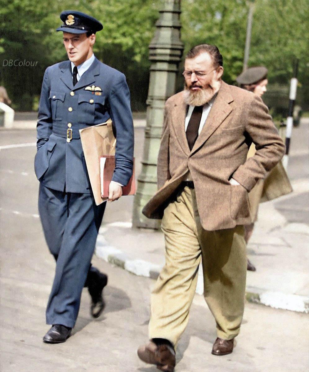 A man in a blue military uniform walks beside an older man with a beard wearing a brown jacket and tan pants on a city sidewalk, both appearing engaged and focused. Trees and a streetlamp are in the background.