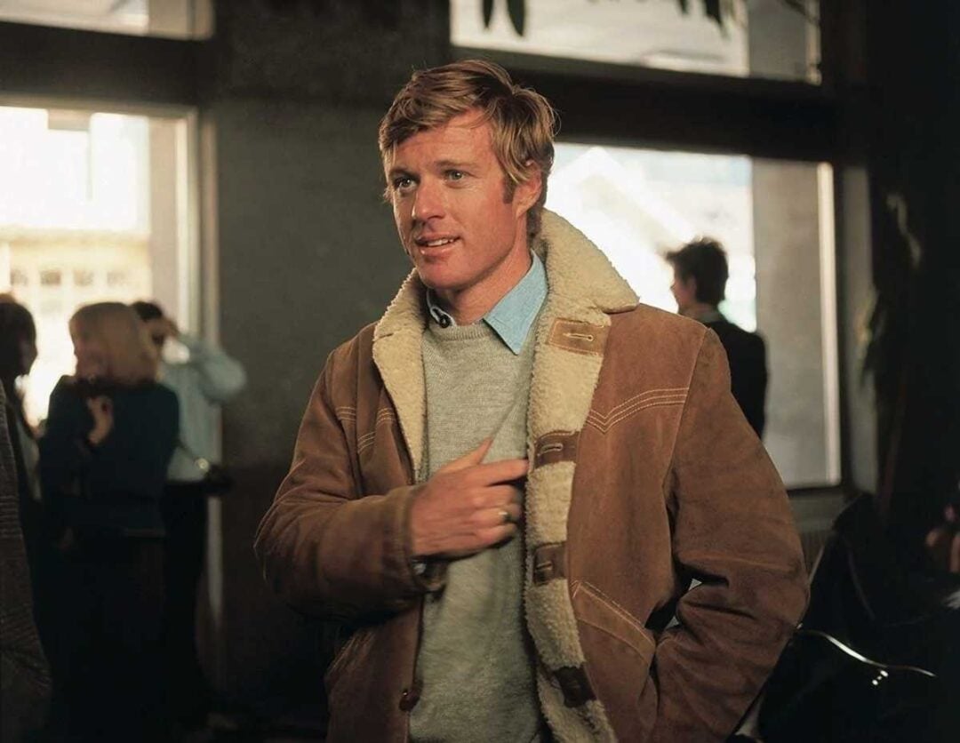 A man with blonde hair, wearing a brown shearling jacket over a gray sweater and collared shirt, stands indoors with people and large windows in the background.