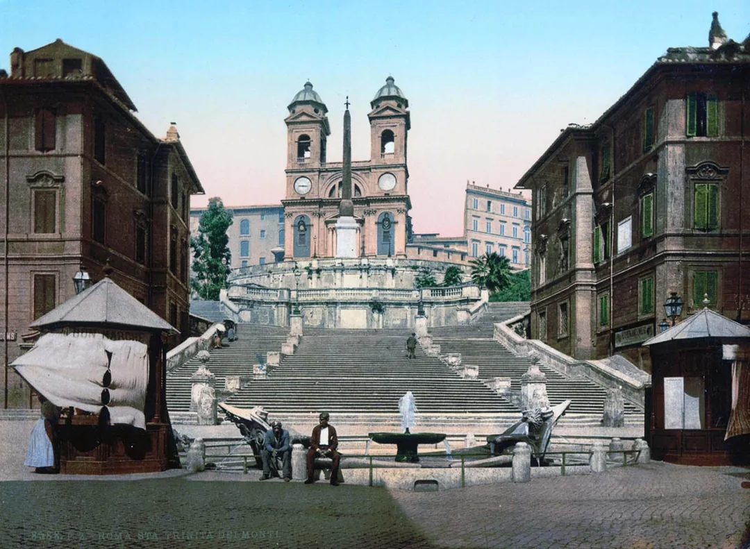 Historic photo of the Spanish Steps in Rome, with people sitting and standing near the base, street vendors, and the Trinità dei Monti church at the top under a clear sky.