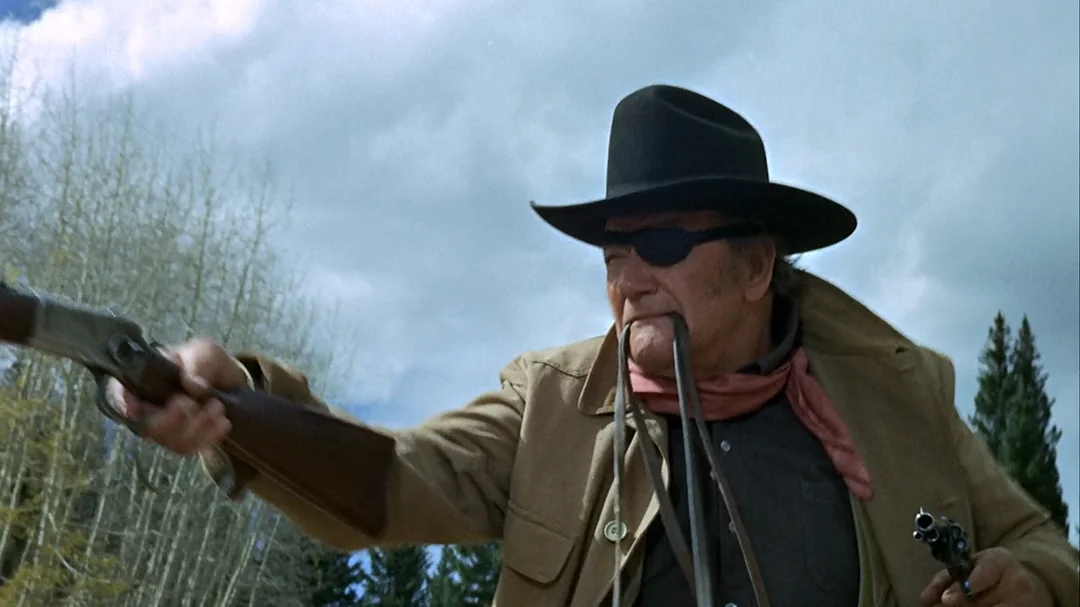 An older man wearing a black hat, eyepatch, brown coat, and red scarf aims a rifle outdoors under a cloudy sky, with trees in the background.