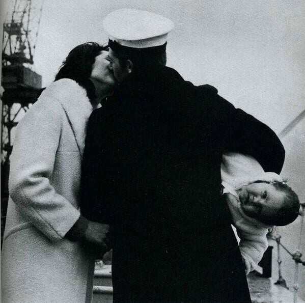 A sailor in uniform kisses a woman, both holding hands, while holding a smiling young child under his arm. Industrial cranes appear in the background on a cloudy day.