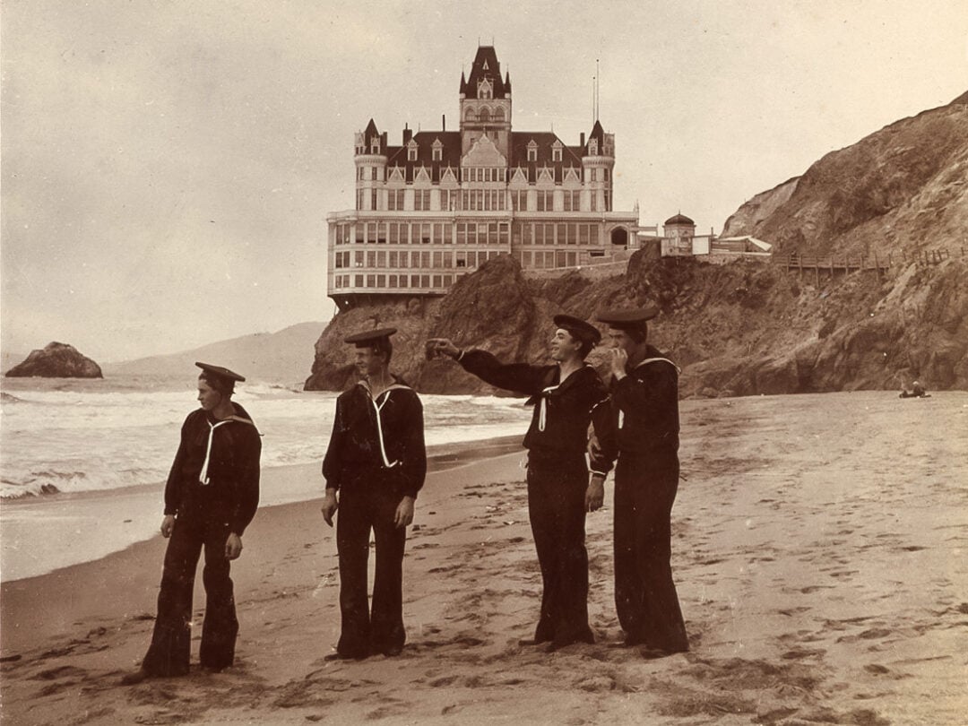 Four sailors in uniform stand on a sandy beach with cliffs behind them. One points toward the ocean. In the background is a large, ornate Victorian-style building perched on a cliff above the waves.