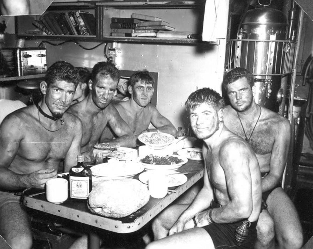Five shirtless men, dirty and sunburned, sit around a small table covered with food and drinks inside a cramped, metal-walled room, possibly a military vessel. They appear tired but are smiling at the camera.