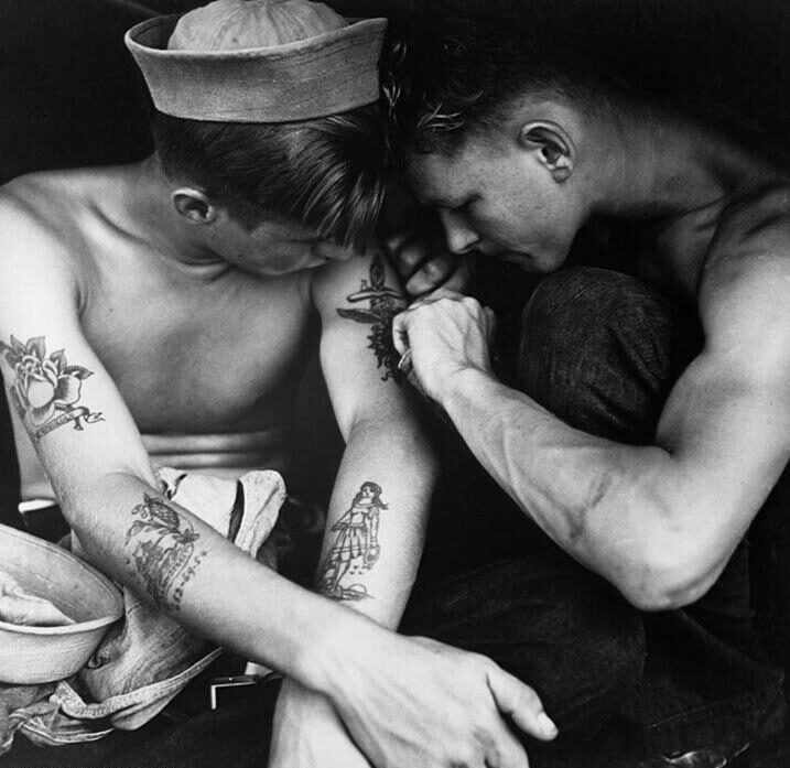 Two young men sit close together; one is tattooing the other's arm. Both are shirtless, with visible tattoos and wearing sailor hats, focused intently on the tattooing process in a black-and-white photograph.