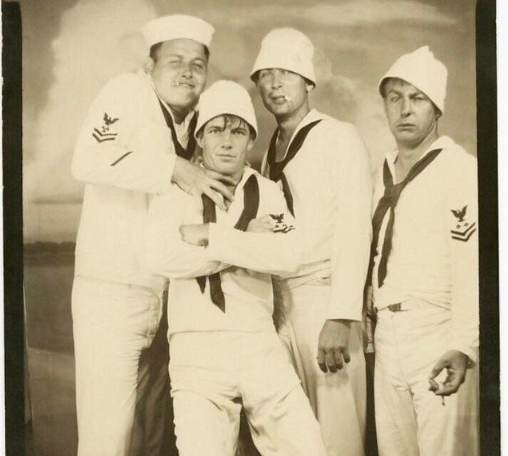 Four men in vintage Navy uniforms pose together in a black-and-white studio photo, with three standing and one crouching. They wear sailor hats and neckerchiefs and have playful, relaxed expressions.