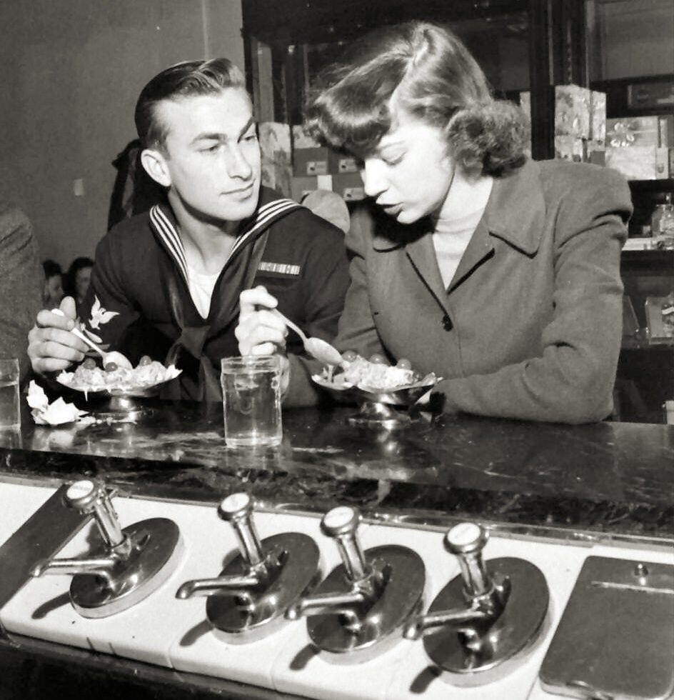 A sailor in uniform and a woman in a coat sit at a diner counter eating dessert together, with soda fountain taps and glasses in the foreground. The image is in black and white and has a LIFE magazine watermark.