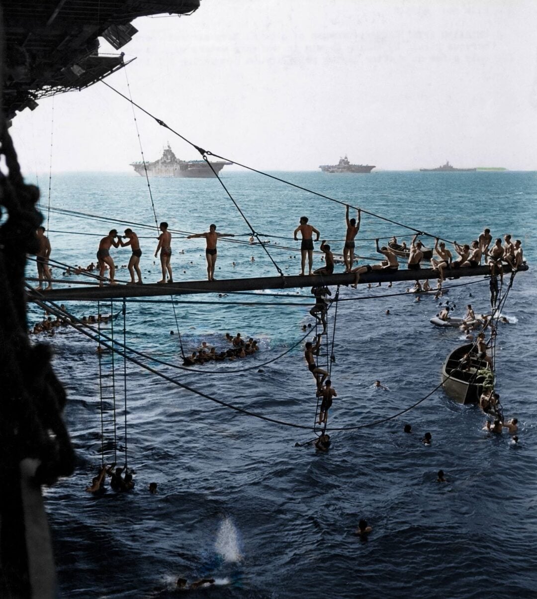Dozens of sailors swim, climb ropes, and jump from a platform extending from a ship into the sea, with other ships visible on the horizon under a clear sky.