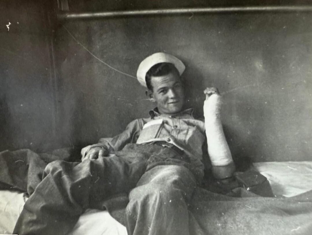 A sailor in uniform reclines on a bed, smiling at the camera, with his right arm in a cast and propped up, suggesting he is recovering from an injury. The background appears to be a simple, metal-walled room.