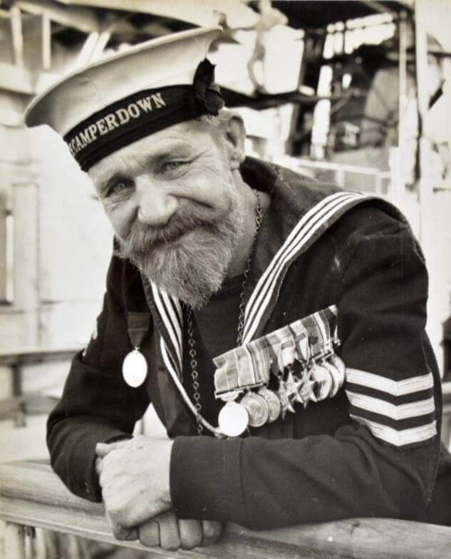 A smiling sailor with a bushy beard wearing a "Camperdown" cap, navy uniform, and multiple medals leans on a railing aboard a ship. The background shows parts of the ship's structure.