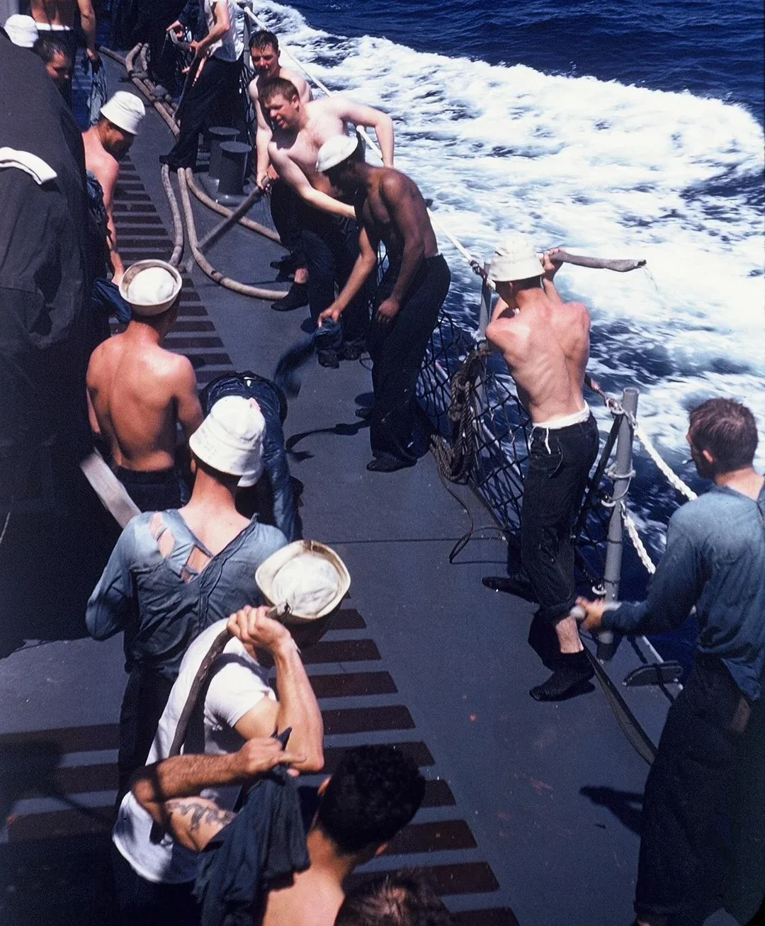 Sailors participate in the 1957 Shellback initiation, "The Gauntlet," on a ship’s deck at sea; some are shirtless, some wear hats, and others hold ropes, with ocean waves visible alongside the ship.