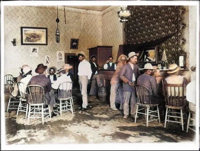 A group of men in hats and vests sit at tables and stand at a wooden bar inside an old-fashioned saloon with patterned wallpaper, framed pictures, and a mirror behind the bar.