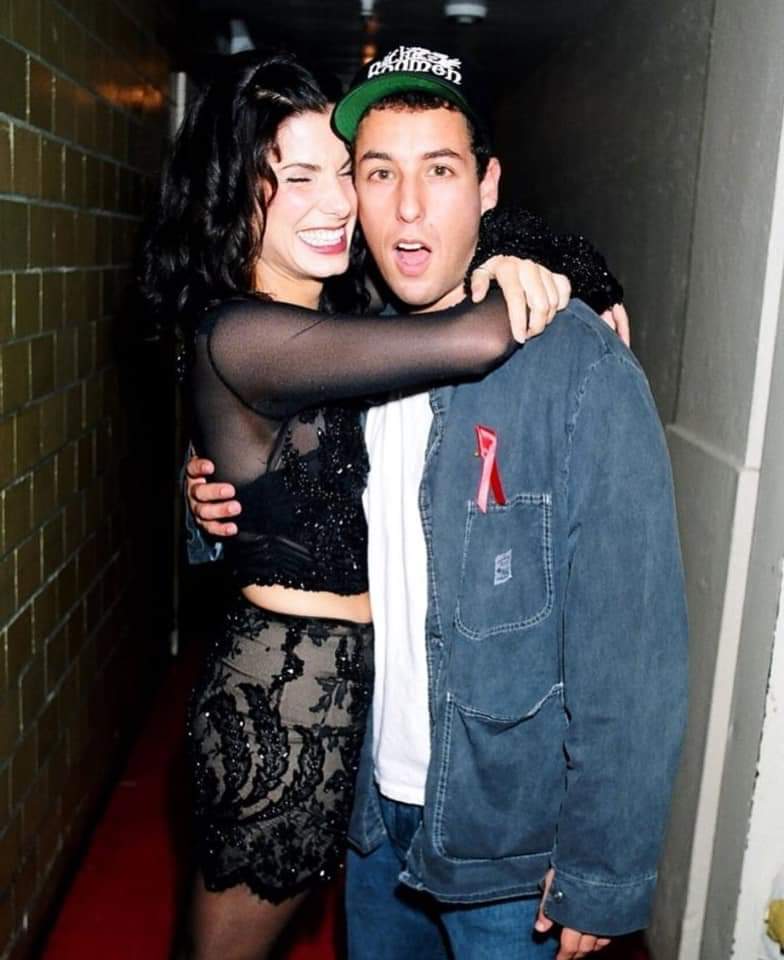 A woman in a black sheer top and patterned skirt hugs a surprised-looking man in a denim jacket and cap. Both are standing in a hallway, smiling at the camera.