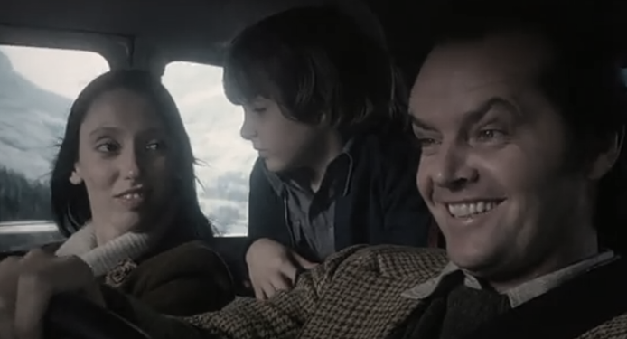 A woman, a boy, and a smiling man are sitting together in a car, with mountains and snow visible through the windows in the background. The man is driving while the boy looks at him and the woman glances back at the boy.