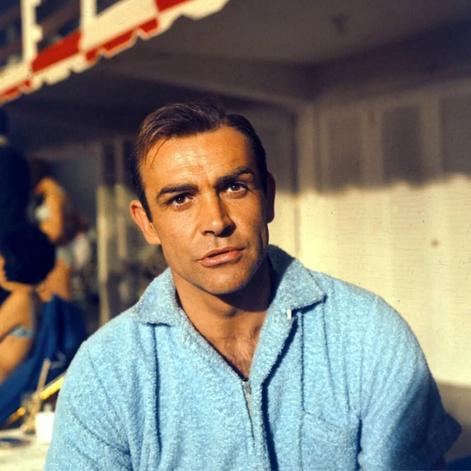 A man with short brown hair, wearing a light blue zip-up terrycloth top, sits outdoors in sunlight. The background shows a woman in blue and red-and-white striped awnings.