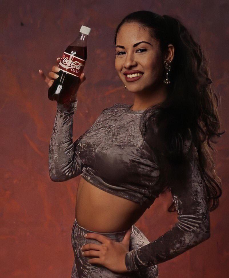 A smiling woman with long dark hair in a high ponytail holds a glass Coca-Cola bottle. She is wearing a shiny, fitted gray velvet crop top and matching pants, standing against a reddish-brown background.