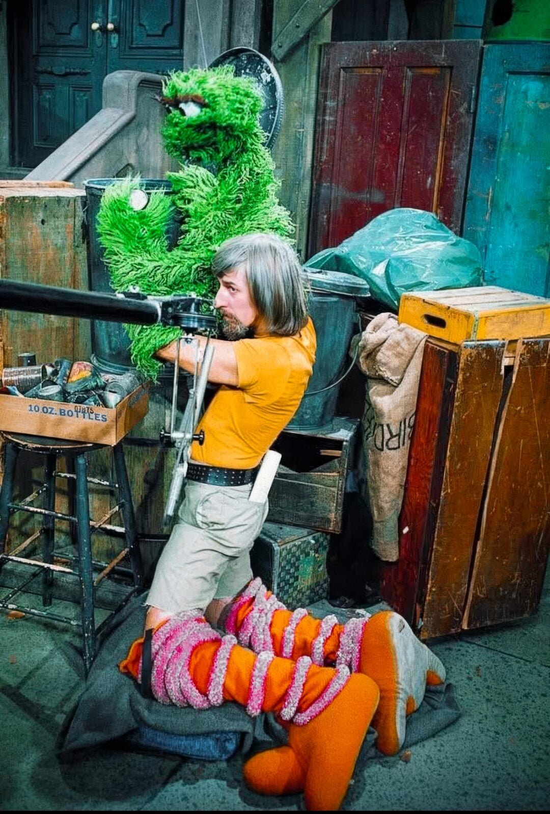 A person kneels on the ground, peering through a large camera. Behind them, the green puppet Oscar the Grouch pops out of a trash can. Bright orange and pink fuzzy legs are in the foreground, suggesting another puppet nearby.