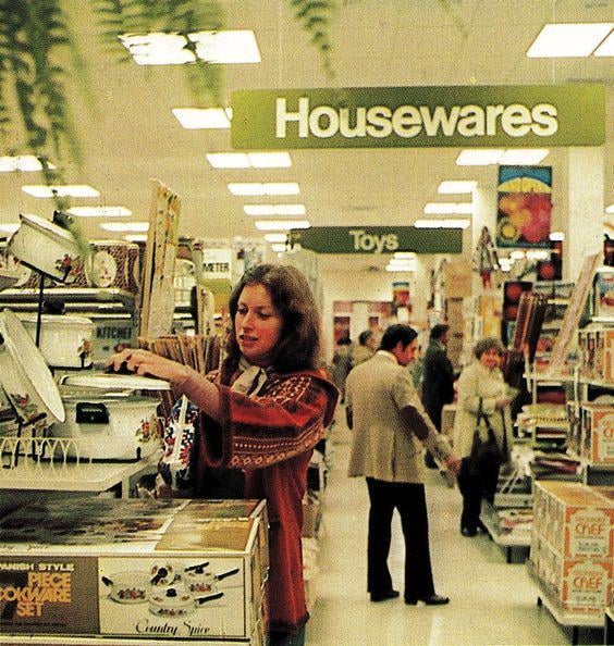 A woman shops in the housewares aisle of a store, surrounded by boxed cookware sets and kitchenware. Other shoppers and an overhead sign labeled "Housewares" are visible in the brightly lit store.
