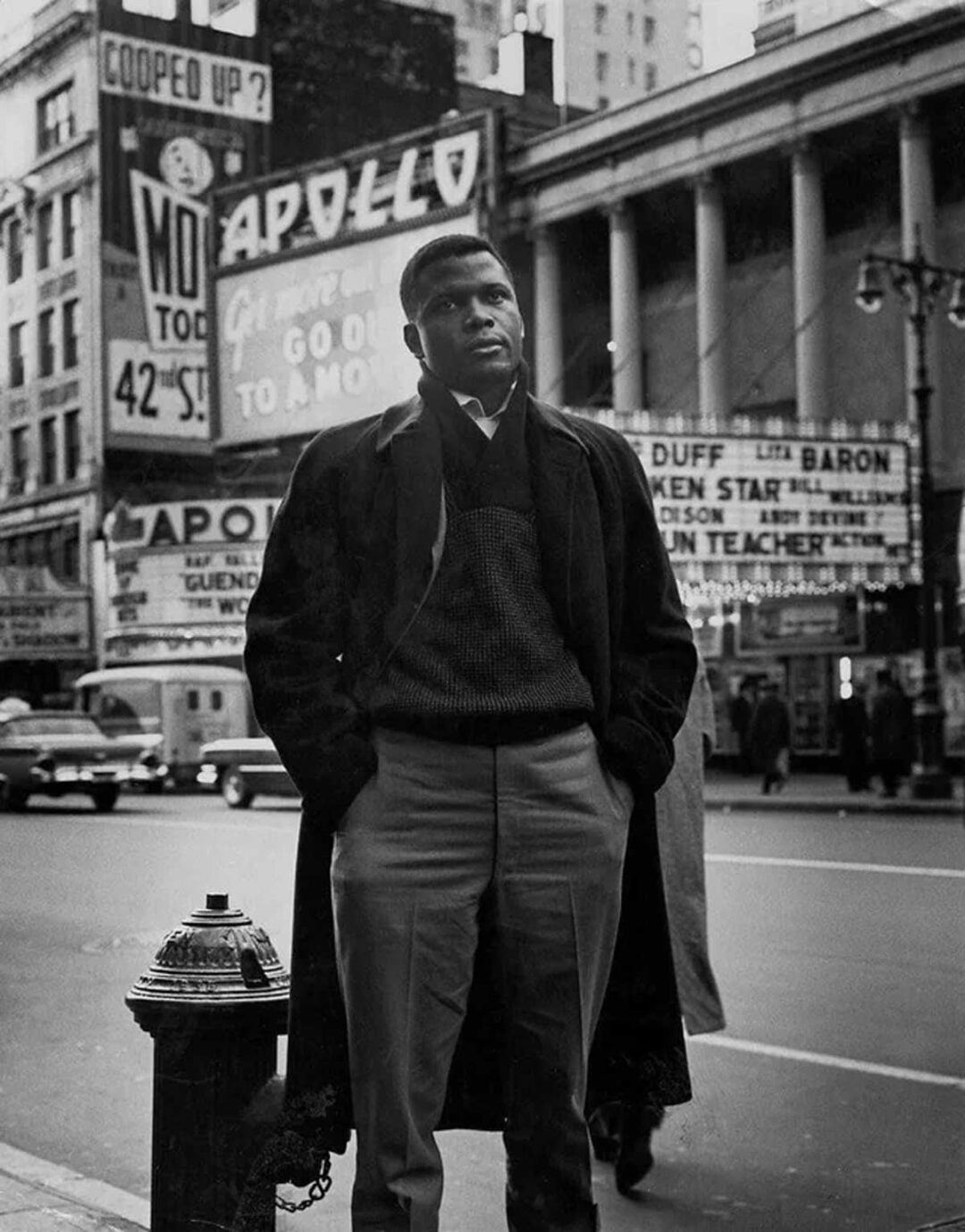 A man wearing a coat stands on a city sidewalk near a fire hydrant. Behind him are 1950s-era cars and the Apollo Theater, with large advertising signs and marquees visible on nearby buildings.