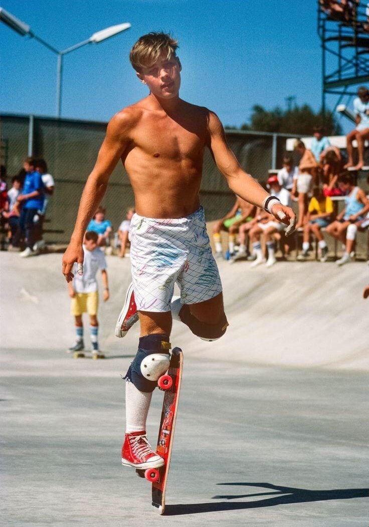 A young man with blond hair, wearing patterned shorts, red high-top sneakers, and kneepads, performs a trick on a skateboard at a skatepark on a sunny day. Spectators watch in the background.