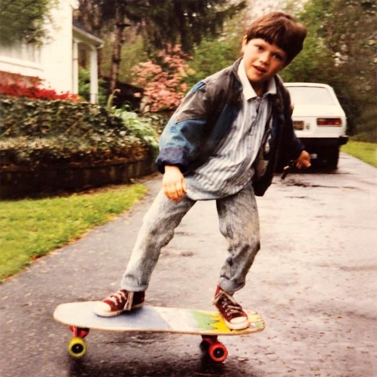 A young boy wearing a jacket, striped shirt, and jeans rides a skateboard on a wet driveway, with one foot on the board and the other off, near a white car and a house surrounded by greenery.