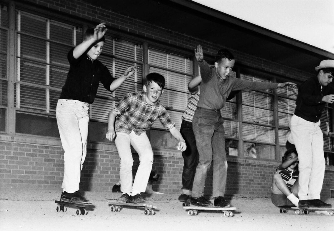 Five boys ride skateboards in front of a brick building with large windows. Four are standing on their boards, arms out for balance, while one sits on his skateboard in the background. The photo is in black and white.