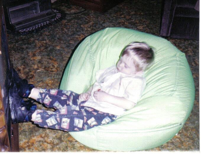 A young child is sleeping in a green bean bag chair, wearing patterned pants, a light shirt, and dark shoes, with feet propped up against a piece of furniture on a carpeted floor.