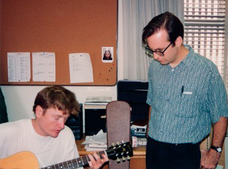 Two men are in an office; one is seated and playing an acoustic guitar, while the other stands beside him watching. Behind them is a corkboard with papers and a small photo pinned to it. Light streams through a window with blinds.
